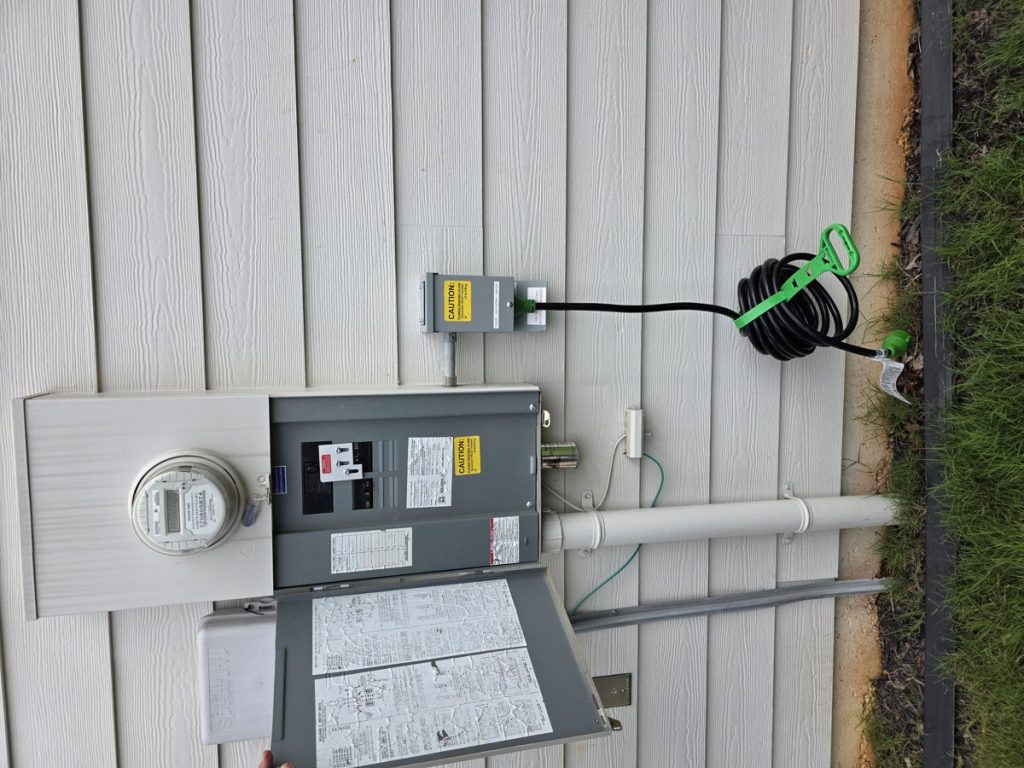 Generator inlet box with a connected generator cord mounted beside a meter-main panel on a home with white siding, installed by Shocking Electric Solutions