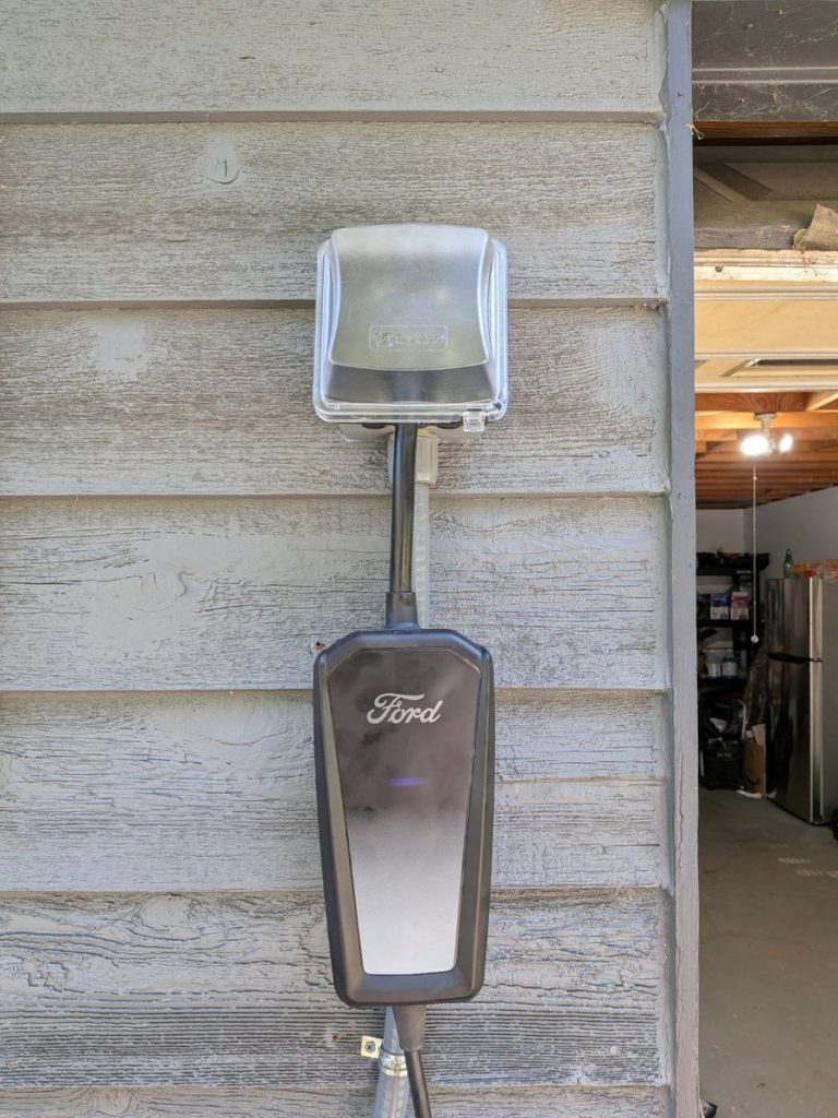 Ford Level 2 home EV charger mounted on gray wood siding beside an open garage, plugged into an outdoor receptacle under a clear in-use weatherproof cover